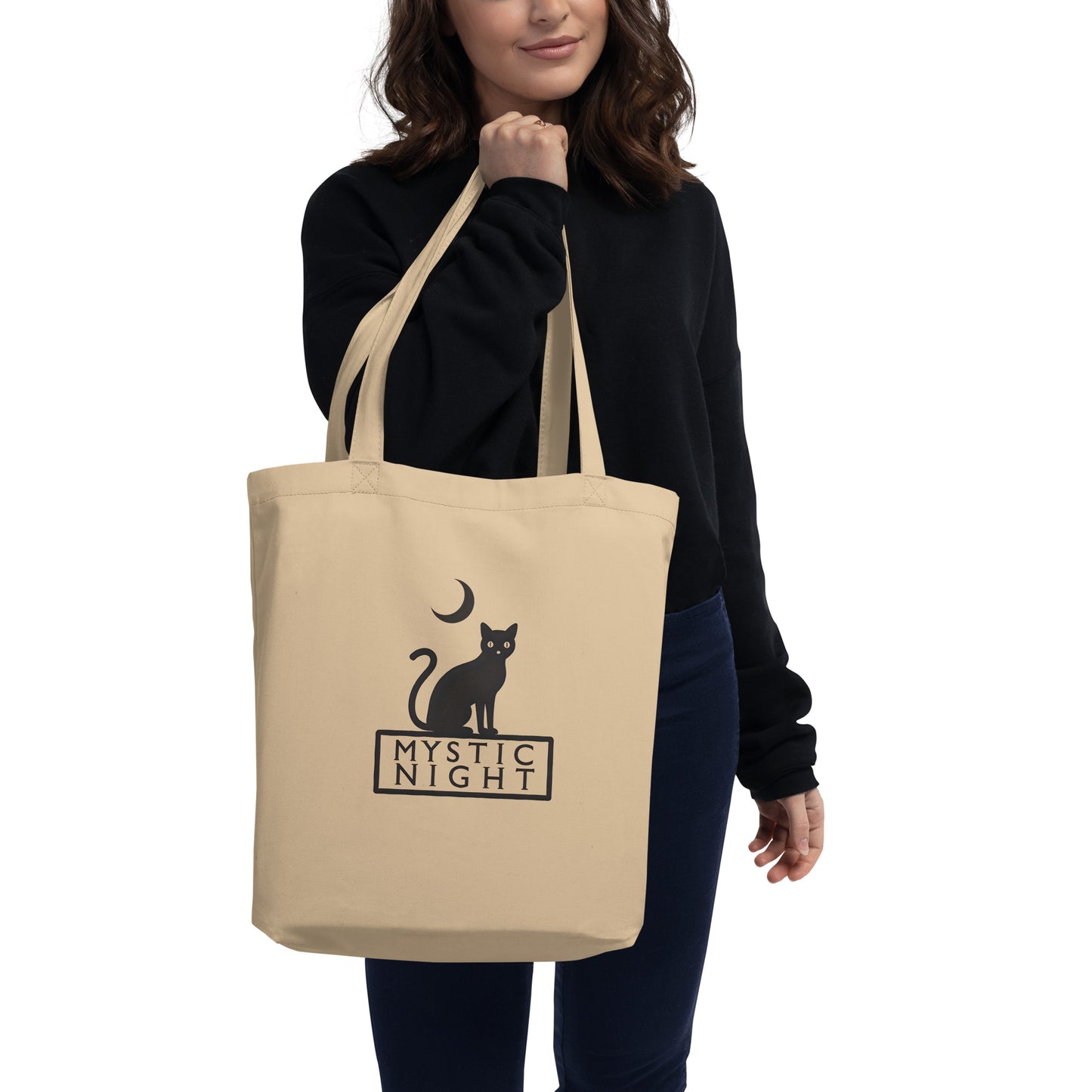 A female model holding a Minimalist oyster color tote bag featuring a black cat sitting under a crescent moon with the text “Mystic Night”.