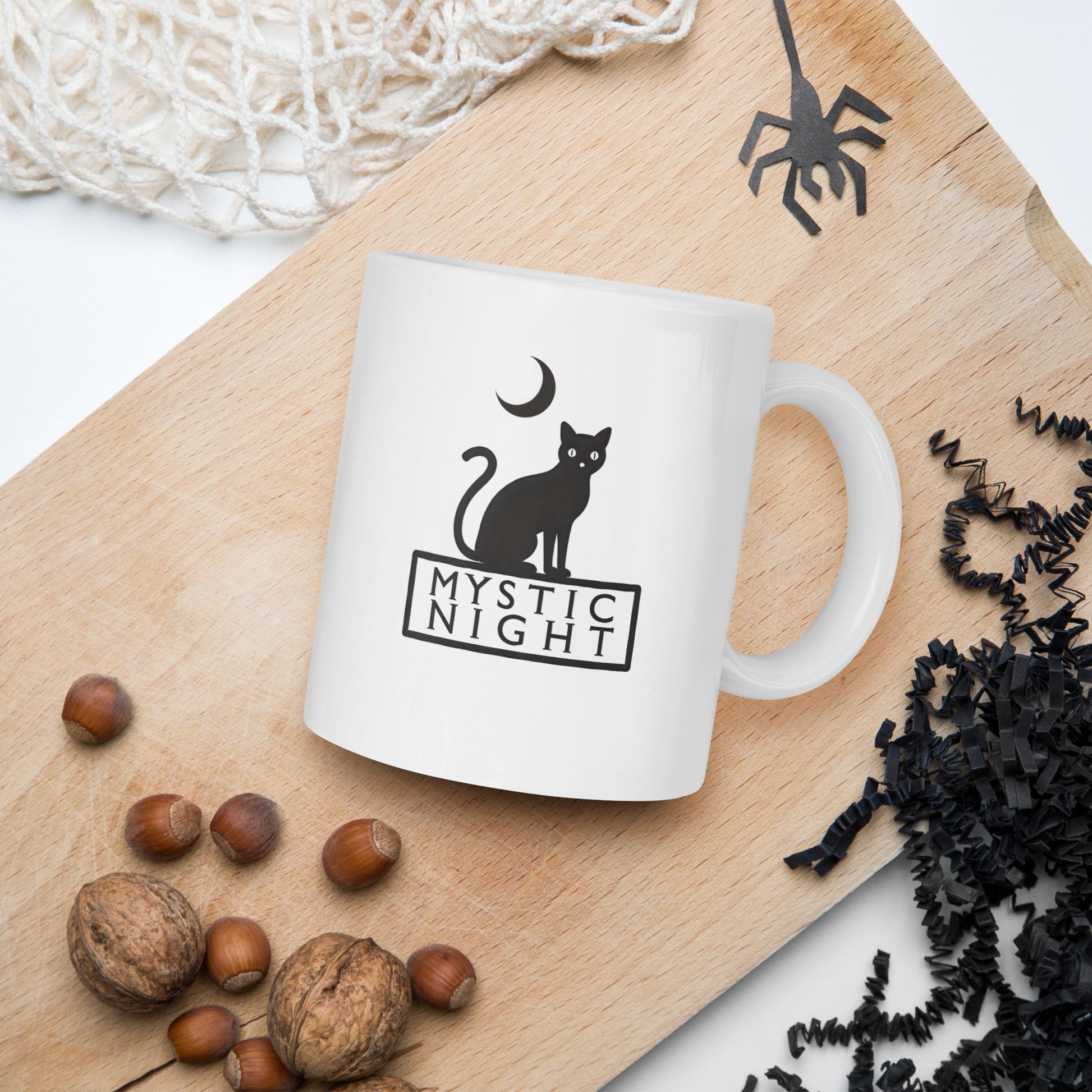 White mug with 'Mystic Night' design featuring a cat and moon on a wooden board with nuts and spider decorations.
